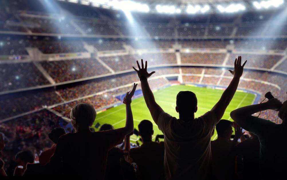 A shot of fans in a stadium, taken from the above the last row of seats. Fans are celebrating as their winning margin bet has landed